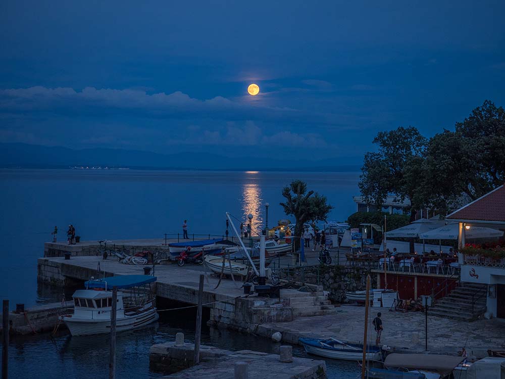 Full moon reflected in the water and a dock with boats and people