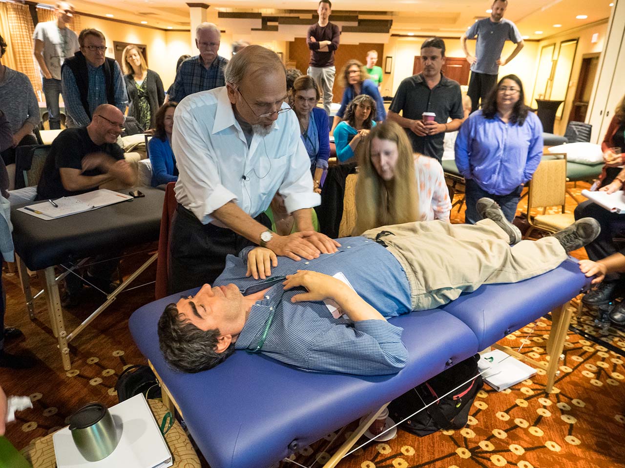 Man lays on a table while another man stands above with two hands on his chest as others look on