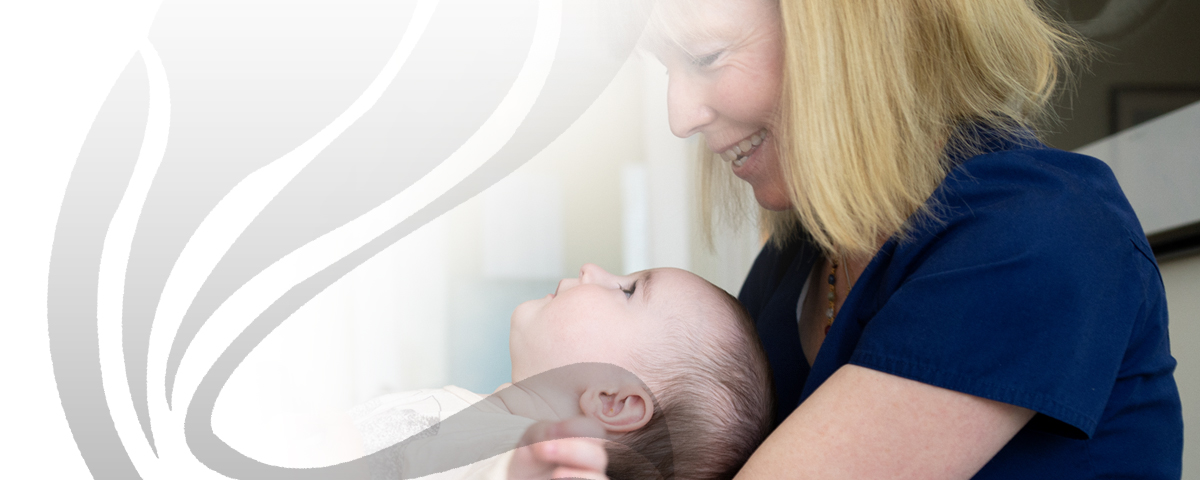 Smiling, a woman holds the back of a baby’s neck as the baby looks up at her