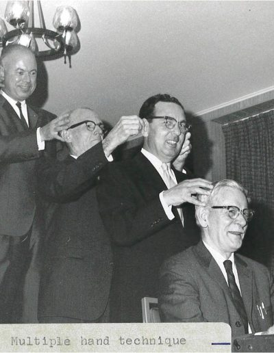 Four men in a line with hands on both sides of the head of the man in front of them